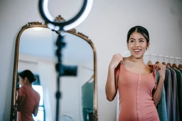 Smiling young ethnic female blogger standing in room and demonstrating pink dress while shooting vlog on smartphone with ring lamp near mirror and wardrobe. UGC creators: UK discovery tools and checks