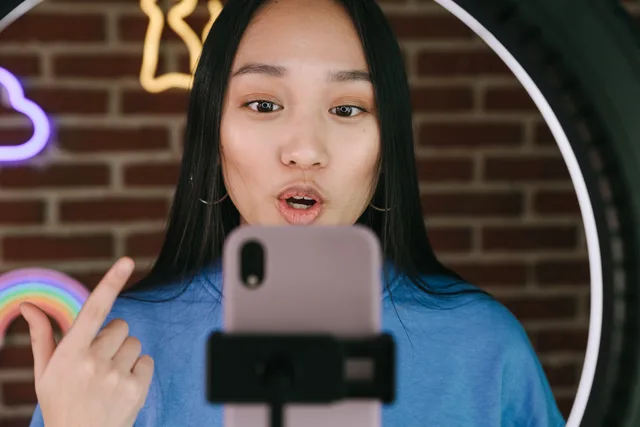 Asian woman vlogging with smartphone and ring light, in a creative indoor setting. UGC marketplaces AI applications: fictional task ideas