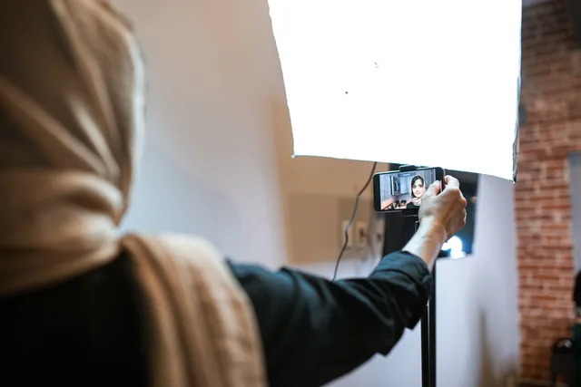 A woman wearing a headscarf is recording a vlog with a smartphone and bright studio light. UGC marketplaces attribution methods for one asset