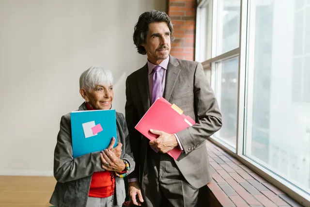 A mature man and woman in business attire stand by office windows holding colorful folders. UGC opportunities treated as buyer problems, with counter-signals and stop rules