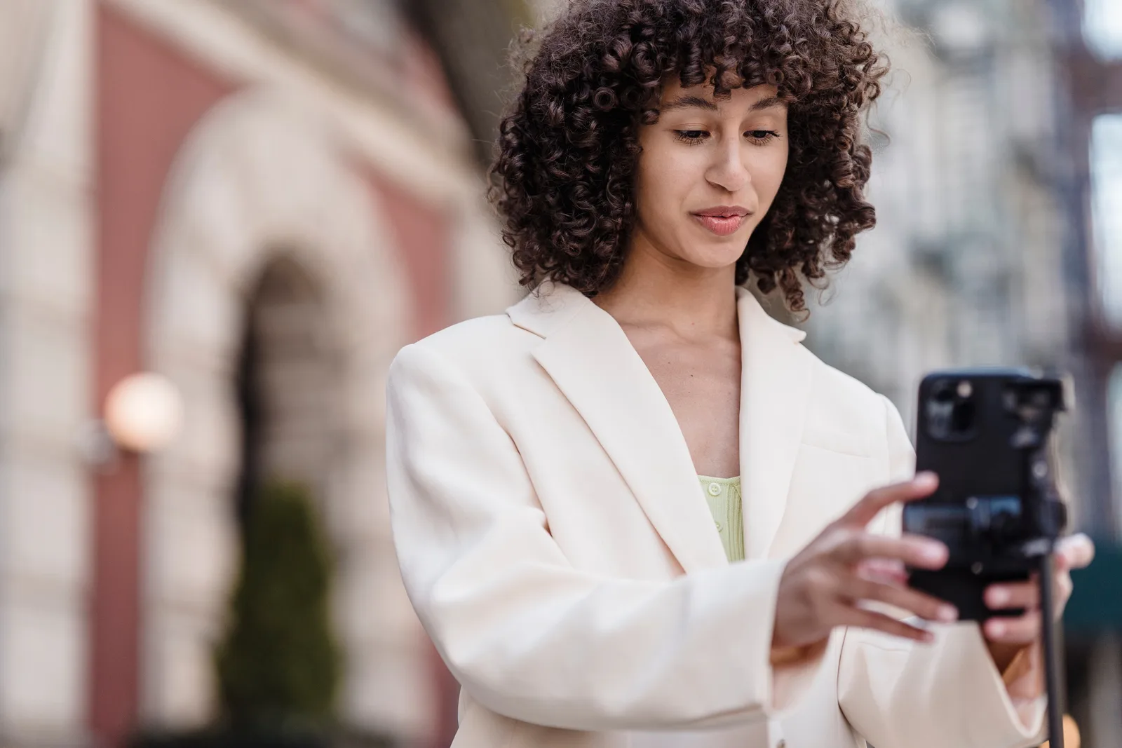 Young ethnic female vlogger with curly hair in jacket preparing cellphone for video record in town