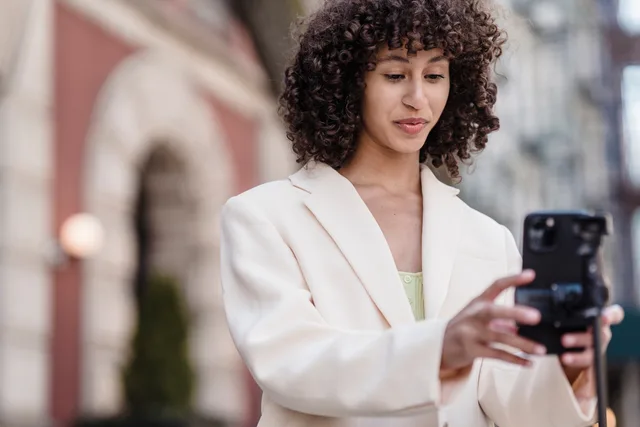 Young ethnic female vlogger with curly hair in jacket preparing cellphone for video record in town. UGC return on investment: the ROI sum, ledger and break-even for one England asset