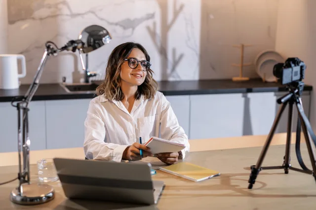 Young woman in glasses working indoors with laptop and camera, smiling while taking notes. Buyer-owned UGC service measures, from admission decision time to exit completeness