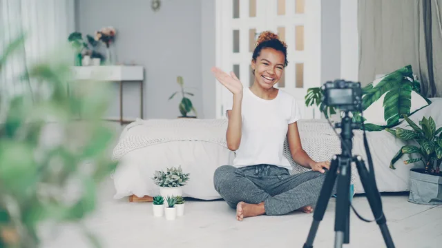 Smiling woman waves at camera while recording a vlog in a stylish bedroom setting. UGC marketplaces target audience: recruiting each role from recent commissions
